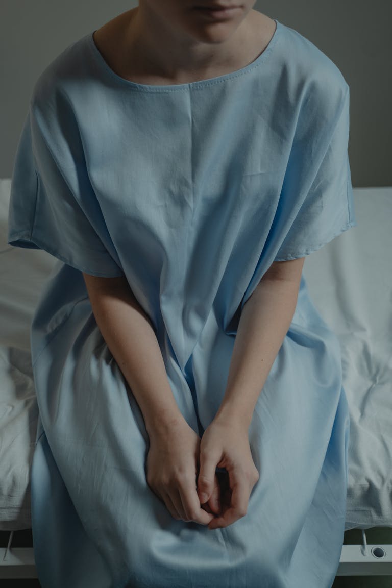 A person sits thoughtfully on a hospital bed wearing a gown, evoking a sense of solitude and contemplation.
